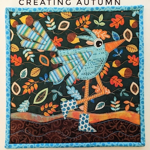 Könnte beinhalten: Ein blauer Vogel mit orangenen Beinen und blauen Stiefeln steht auf einem braunen Hintergrund mit einem Muster aus grünen, orangen und braunen Blättern. Der Text "CREATING AUTUMN" befindet sich oben im Bild.