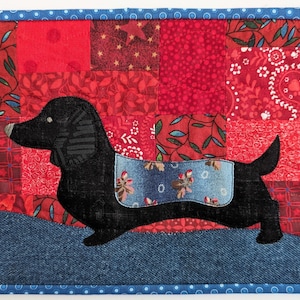 May include: A black dachshund applique on a red and blue patchwork quilt. The quilt is made of various fabrics, including denim, paisley, and floral prints.