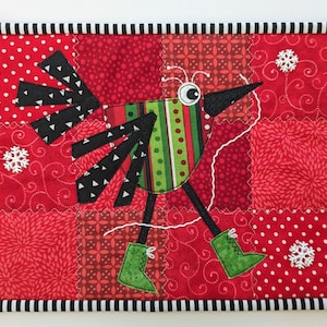 Könnte beinhalten: Eine rote, grüne und schwarze Patchwork-Decke mit einem skurrilen Vogeldesign. Der Vogel hat einen langen, dünnen Schnabel, große Augen und trägt grüne Stiefel. Die Decke ist mit Schneeflocken verziert.