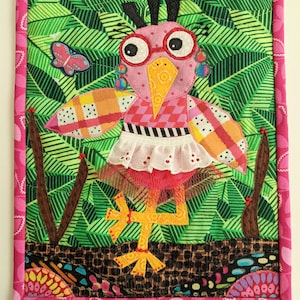 Könnte beinhalten: Ein rosa-gelber Vogel mit großen Augen und Brille steht auf einem Bein in einem grünen Blattwerk. Der Vogel trägt ein weißes Kleid mit einem rosa-orangenen gemusterten Oberteil. Der Vogel hat schwarze Haare und rosa Ohrringe. Der Hintergrund ist ein grünes und braunes Patchwork-Design. Der Text "CREATING MIA" befindet sich am unteren Rand des Bildes.
