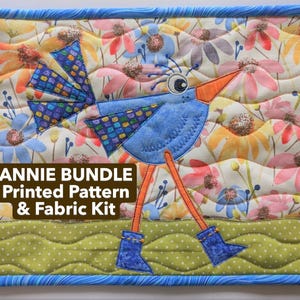 May include: A quilted fabric kit featuring a whimsical blue bird design with an orange beak and legs, and blue boots. The background has a floral pattern with a green wavy border and a blue patterned frame. The text "ANNIE BUNDLE Printed Pattern & Fabric Kit" is displayed.