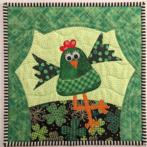 May include: A green quilted fabric wall hanging featuring a green chicken with orange legs and black shamrock wings. The chicken is standing on a black background with green shamrocks.