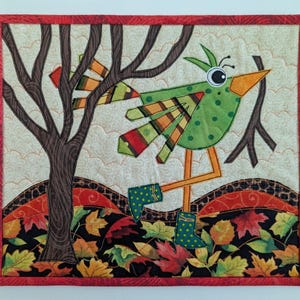 Könnte beinhalten: Ein gestepptes Wandbehang mit einem skurrilen grünen Vogel mit orangefarbenen Beinen und einem gelben Schnabel, der blaue gepunktete Stiefel trägt. Der Vogel ist vor dem Hintergrund eines Baumes, Herbstlaub und einem roten Rand platziert. Das Kunstwerk ist eine farbenfrohe Darstellung des Herbstes.