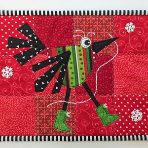 May include: A quilted bird design on a red patchwork background. The bird has a black beak, black and white wings, and green and red striped body. It wears green boots and is surrounded by white snowflakes. The border is black and white striped.