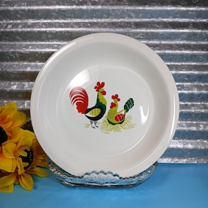 May include: White ceramic plate with a rooster and hen design. The rooster has a red comb and tail, with green and black body. The hen has a red comb and green and yellow body. The plate is on a clear glass stand.