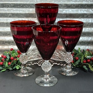 May include: A set of four deep red glass goblets with clear, textured, spherical stems and circular bases. The bowls of the glasses feature a smooth, translucent crimson finish, while the stems have a distinct hobnail or diamond-cut pattern.