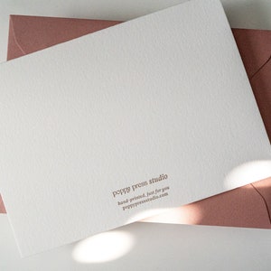 Peut inclure: Une carte blanche avec une surface textur&eacute;e et le texte "poppy press studio" en lettres brunes. Le texte ci-dessous indique "hand-printed, just for you" et "poppy press studio.com". La carte est sur une enveloppe rose.