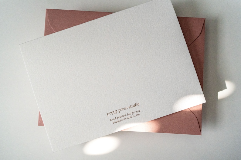 Peut inclure: Une carte blanche avec une surface textur&eacute;e et le texte "poppy press studio" en lettres brunes. Le texte ci-dessous indique "hand-printed, just for you" et "poppy press studio.com". La carte est sur une enveloppe rose.