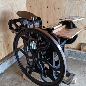 Peut inclure: Une presse d'impression vintage noire avec une grande roue et une plateforme en bois. La presse est sur une base en bois et poss&egrave;de un levier en m&eacute;tal.