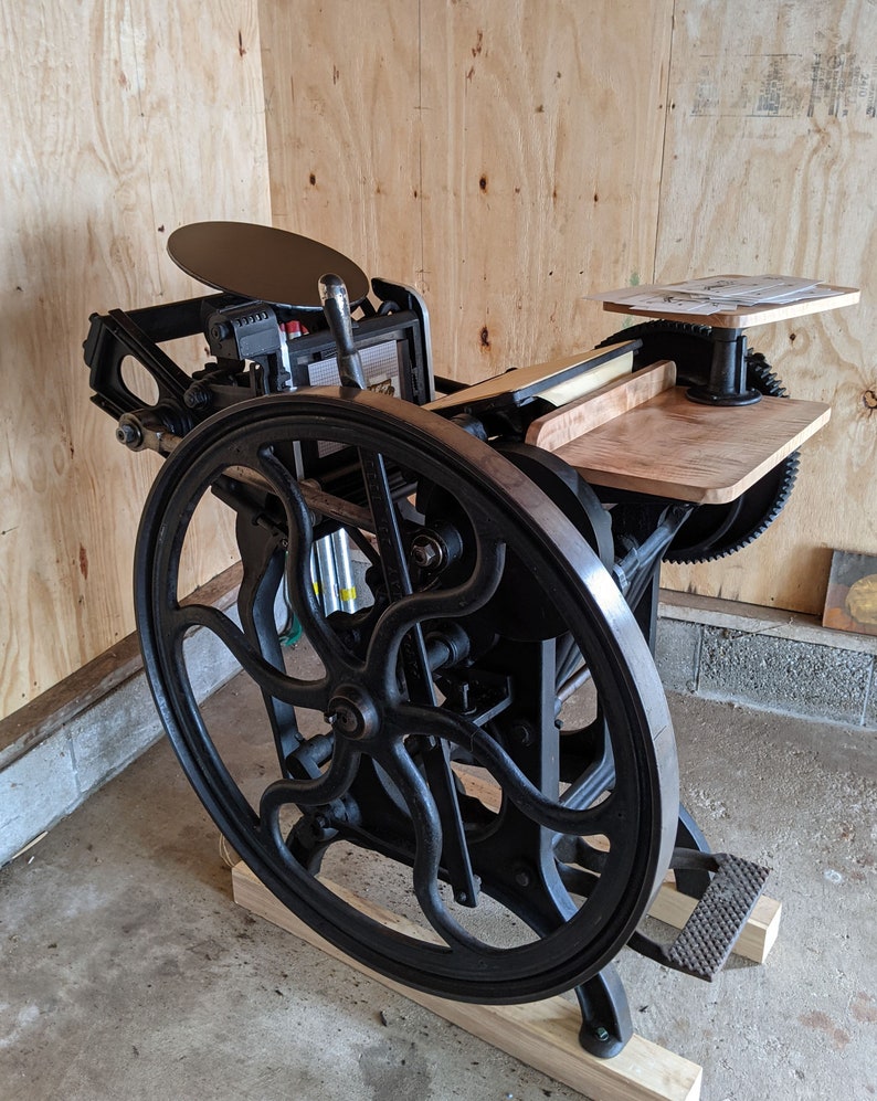 Peut inclure: Une presse d'impression vintage noire avec une grande roue et une plateforme en bois. La presse est sur une base en bois et poss&egrave;de un levier en m&eacute;tal.