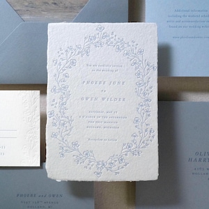 May include: Wedding stationery set featuring a central invitation with floral wreath design and text. Includes RSVP card, envelopes, and additional informational cards in shades of blue and white.