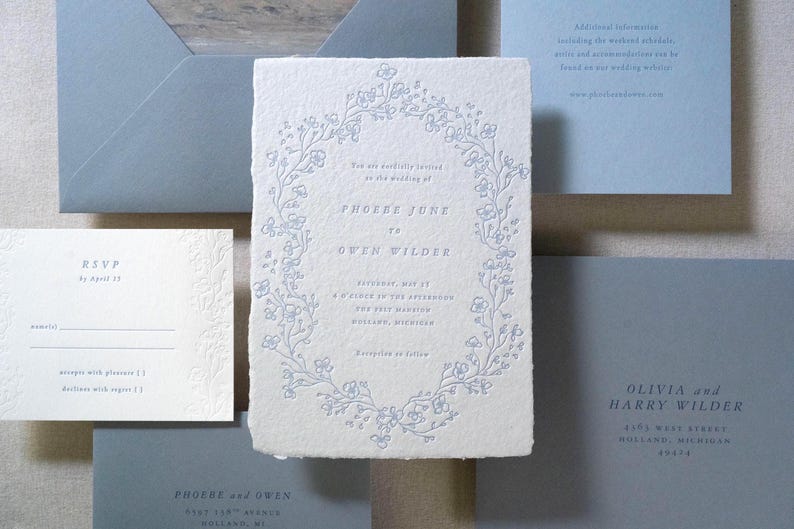 May include: Wedding stationery set featuring a central invitation with floral wreath design and text. Includes RSVP card, envelopes, and additional informational cards in shades of blue and white.