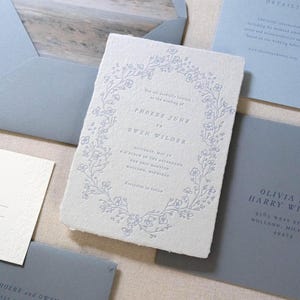 May include: Wedding invitation suite featuring a central card with floral wreath design and text. Includes RSVP, details, and envelope in shades of blue and white. The invitation text reads "Phoebe Jure to Owen Wilder".