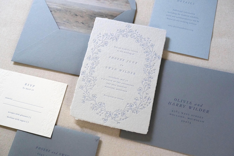 May include: Wedding invitation suite featuring a central card with floral wreath design and text. Includes RSVP, details, and envelope in shades of blue and white. The invitation text reads "Phoebe Jure to Owen Wilder".
