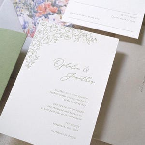 以下が含まれることがあります： 結婚式の招待状セット。白い招待状カードには花のアクセントと「Ophelia & Jonathan」の名前が書かれています。追加のカードには、緑色の詳細カードと灰色の封筒が含まれています。