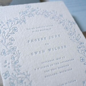 May include: A light blue wedding invitation with a floral border and text. The invitation features the names "PHOEBE JUNE" and "OWEN WILDER", along with the wedding date and location. The paper has a textured, handmade appearance.