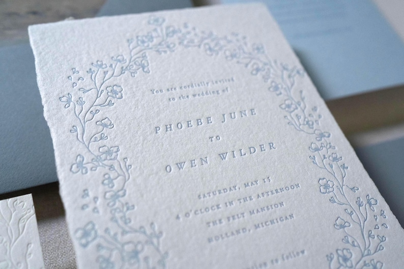 May include: A light blue wedding invitation with a floral border and text. The invitation features the names "PHOEBE JUNE" and "OWEN WILDER", along with the wedding date and location. The paper has a textured, handmade appearance.