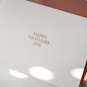 Peut inclure: Une carte blanche avec le texte "Happy Mother's Day" en lettres dor&eacute;es. La carte est pos&eacute;e sur une surface blanche avec une enveloppe rose derri&egrave;re.