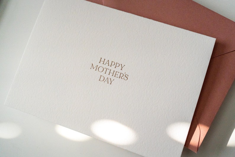 Peut inclure: Une carte blanche avec le texte "Happy Mother's Day" en lettres dor&eacute;es. La carte est pos&eacute;e sur une surface blanche avec une enveloppe rose derri&egrave;re.