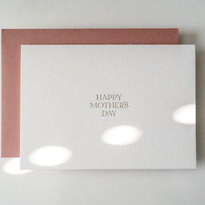 Peut inclure: Une carte blanche avec une enveloppe rose. La carte porte le texte "Happy Mother's Day" imprim&eacute; en brun clair.