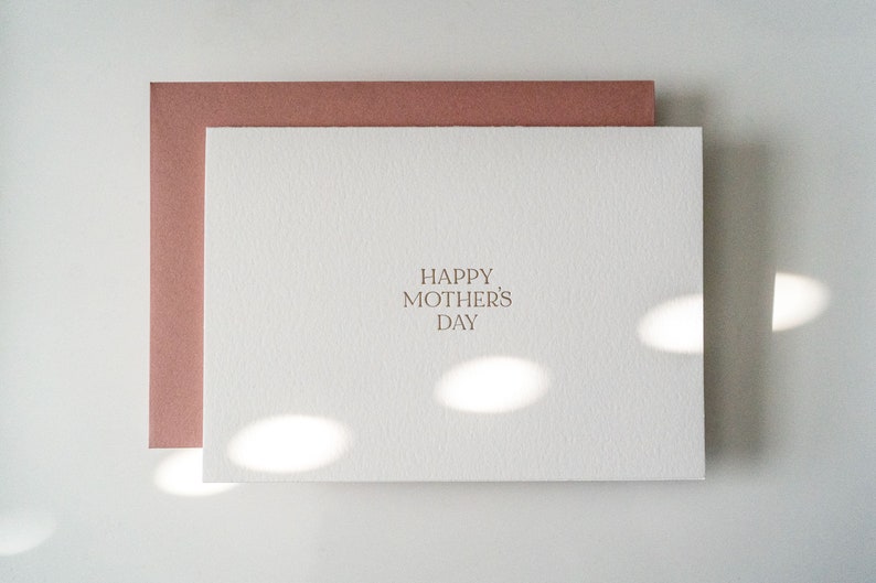 Peut inclure: Une carte blanche avec une enveloppe rose. La carte porte le texte "Happy Mother's Day" imprim&eacute; en brun clair.