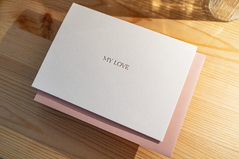 May include: A white card with the text "MY LOVE" in a simple font, layered over a pink envelope. The stationery is on a light wood surface, with soft lighting.