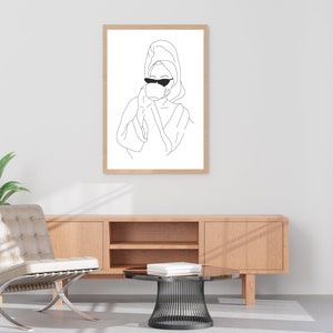 May include: Line art print of a person wearing a towel on their head and sunglasses, drinking from a cup. The print is framed and hanging on a white wall.