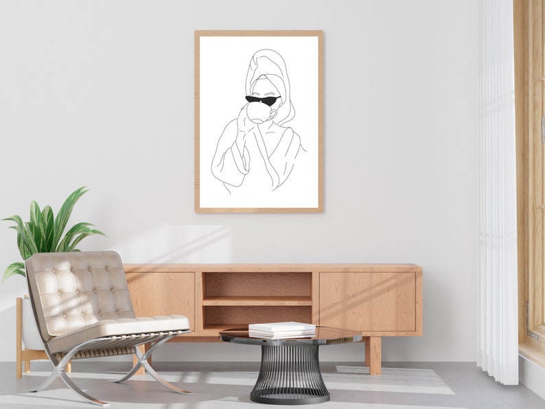 May include: Line art print of a person wearing a towel on their head and sunglasses, drinking from a cup. The print is framed and hanging on a white wall.