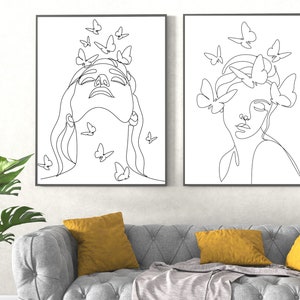 May include: Two black and white line drawings of women's faces with butterflies around them. The drawings are framed and hanging on a white wall above a gray couch with yellow throw pillows.