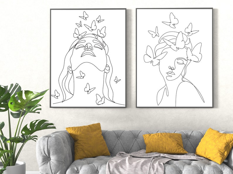 May include: Two black and white line drawings of women's faces with butterflies around them. The drawings are framed and hanging on a white wall above a gray couch with yellow throw pillows.
