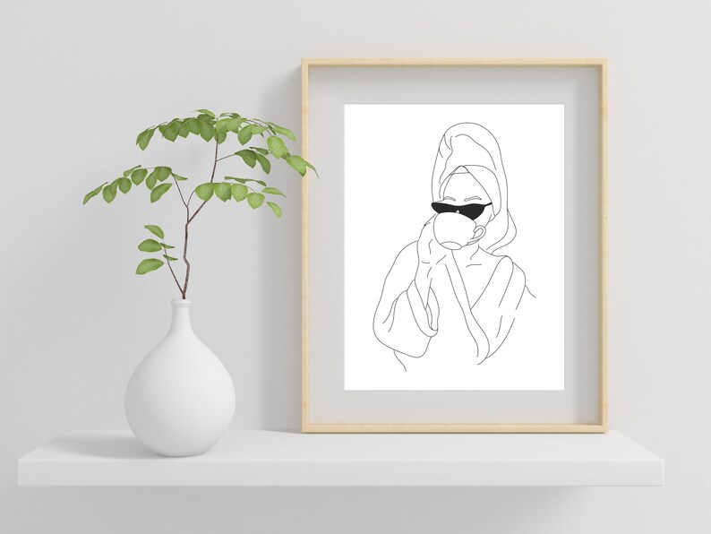 May include: Line art drawing of a person wearing a bathrobe and sunglasses, holding a cup of coffee or tea. The drawing is in black and white and is framed in a light wood frame.