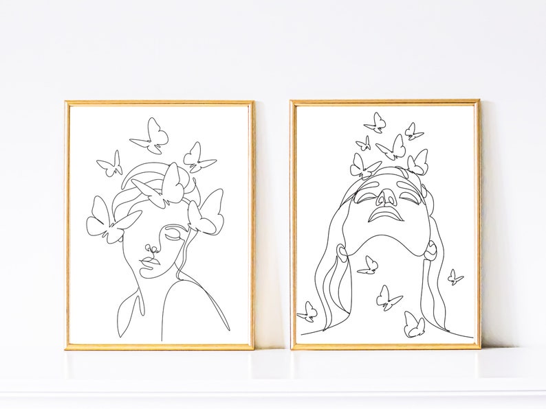 May include: Two black and white line art prints of women's faces with butterflies surrounding them. The prints are framed in gold.
