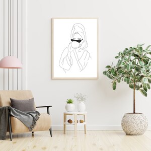 May include: A black and white line drawing of a person wearing a bathrobe and sunglasses, holding a cup. The drawing is framed and hanging on a white wall. A potted plant and a chair are in the foreground.