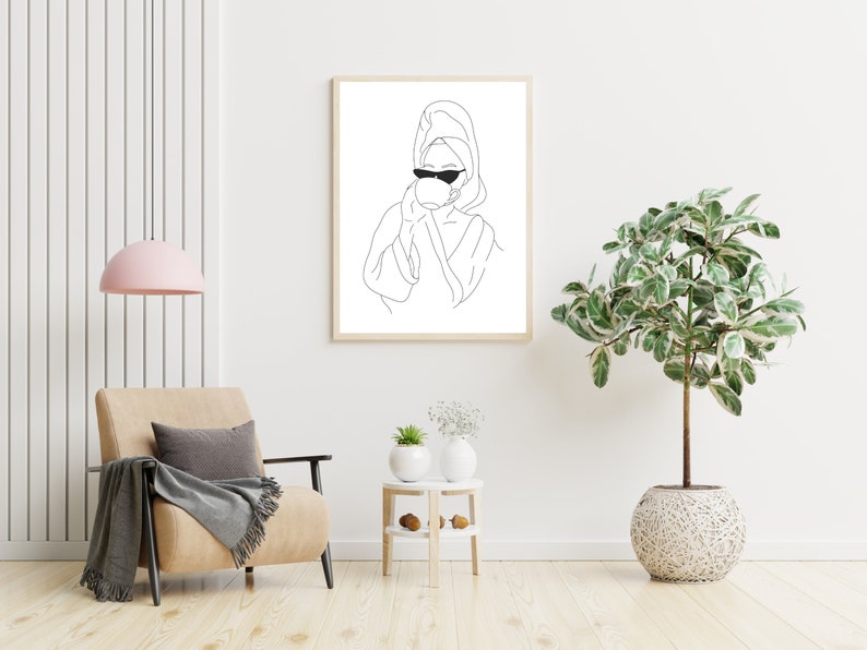 May include: A black and white line drawing of a person wearing a bathrobe and sunglasses, holding a cup. The drawing is framed and hanging on a white wall. A potted plant and a chair are in the foreground.