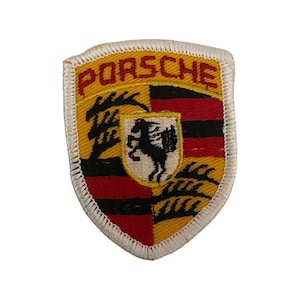 May include: A shield-shaped embroidered patch featuring the Porsche logo in red text at the top. The design includes a central crest with a black horse on a cream background, surrounded by alternating yellow and red-black striped sections.