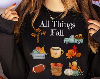 May include: Black sweatshirt with the text "All Things Fall" in white. The design features a variety of fall-themed illustrations, including a truck, pumpkins, leaves, a coffee mug, and a football.