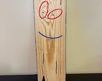 May include: A rectangular wooden board with a natural light brown wood grain finish. The board features a hand-drawn face consisting of two red oval eyes with blue dots, a curved blue line for a mouth, and two vertical black lines at the bottom.