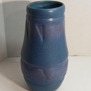 May include: A matte blue ceramic vase with a tapered cylindrical shape. The surface features subtle embossed geometric patterns, including triangular indentations near the rim and a repeating scalloped motif around the lower section of the body.