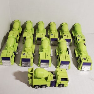 May include: A collection of ten green toy cement mixer trucks. The trucks are all the same design, with a green cab and a rotating cement drum. The trucks are arranged in a row, with the front of each truck facing the viewer.