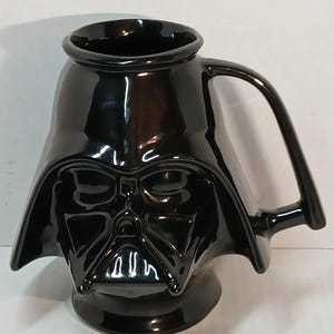 May include: A black ceramic mug shaped like Darth Vader's helmet from Star Wars. The mug has a glossy finish, a handle, and a detailed depiction of the character's face. The mug is designed for beverages.