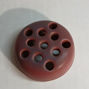 May include: A circular, reddish-brown ceramic flower frog featuring a domed top with ten evenly spaced, circular perforations for arranging stems. The surface has a smooth, matte finish with subtle darker shading around the holes.
