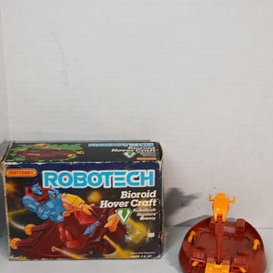 Puede incluir: Juguete vintage Matchbox Robotech Bioroid Hover Craft con caja original. La caja presenta una colorida ilustración del juguete en acción. El juguete en sí es un aerodeslizador rojo y naranja.