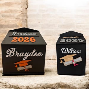 K&ouml;nnte beinhalten: Zwei schwarze Andenkenboxen zur Graduierung. Die gr&ouml;&szlig;ere Box hat die Aufschrift "Graduate 2026" und "Brayden" in wei&szlig;er und orangefarbener Schrift, mit einer Grafik von Abschlusskappe und Diplom. Die kleinere Box hat die Aufschrift "Graduate 2025" und "William" in wei&szlig;er Schrift, mit einer Grafik von Abschlusskappe und Diplom.