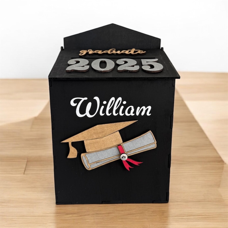 K&ouml;nnte beinhalten: Eine schwarze Holz-Abschlussbox mit dem Namen "William" in wei&szlig;er Schrift. Die Box zeigt ein Design mit Abschlusskappe und Diplom, mit dem Wort "graduate" und dem Jahr "2025" in Holzbuchstaben oben. Die Box steht auf einer Holzoberfl&auml;che.