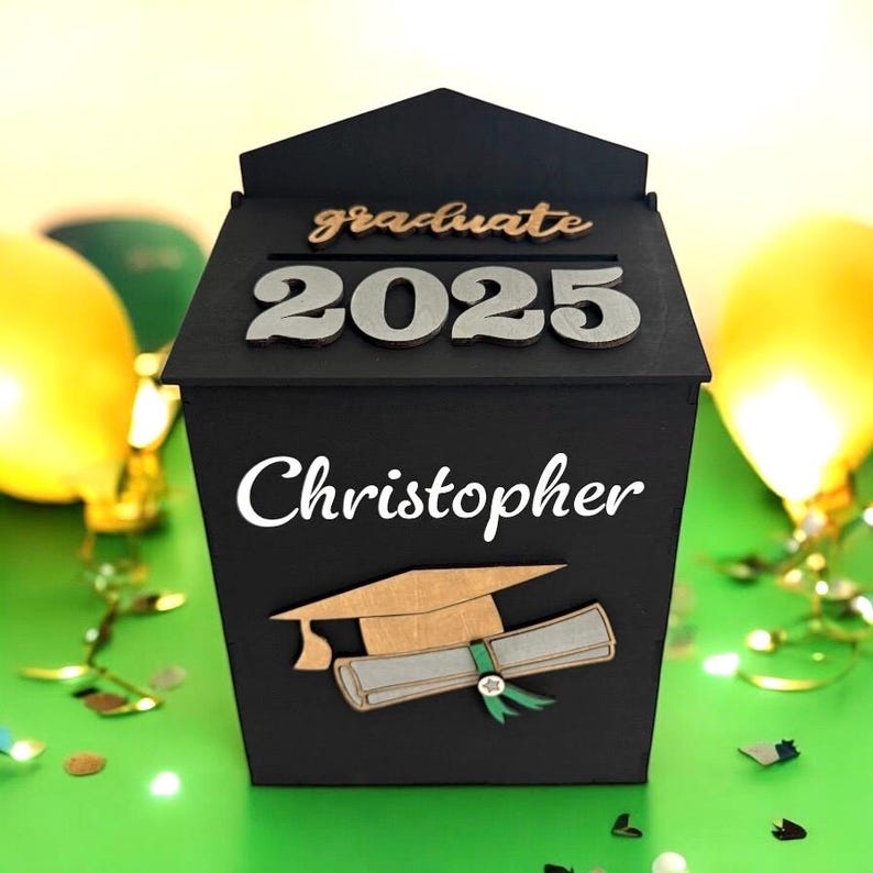 K&ouml;nnte beinhalten: Eine schwarze Holz-Abschlussbox mit dem Namen "Christopher" in wei&szlig;er Schrift. Die Box zeigt "graduate 2025" in silbernen Buchstaben und ein Design mit Abschlusskappe und Diplom. Gelbe Luftballons und Konfetti sind im Hintergrund.
