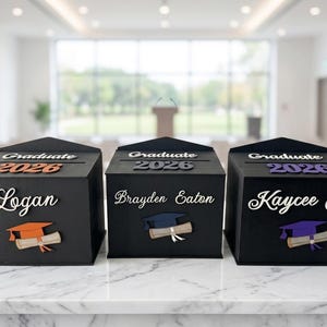 K&ouml;nnte beinhalten: Drei schwarze Abschlussboxen mit personalisierten Namen und dem Jahr 2026. Jede Box zeigt ein Design mit Abschlusskappe und Diplom. Die Boxen werden auf einer wei&szlig;en Marmoroberfl&auml;che pr&auml;sentiert.