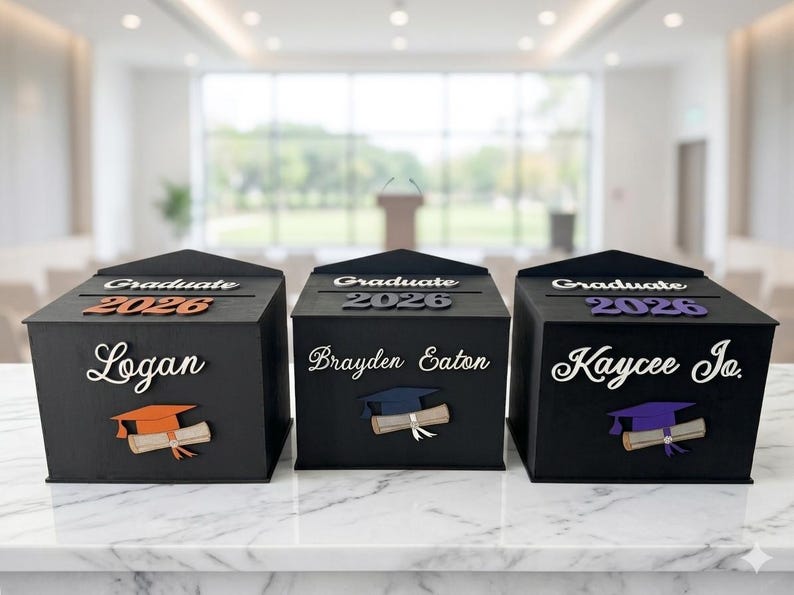 K&ouml;nnte beinhalten: Drei schwarze Abschlussboxen mit personalisierten Namen und dem Jahr 2026. Jede Box zeigt ein Design mit Abschlusskappe und Diplom. Die Boxen werden auf einer wei&szlig;en Marmoroberfl&auml;che pr&auml;sentiert.