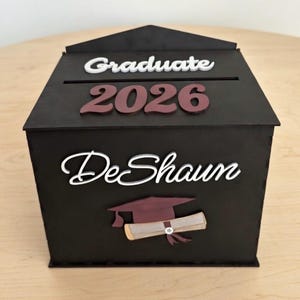 K&ouml;nnte beinhalten: Eine schwarze Holz-Abschlussbox mit einem Schlitz oben. Die Box zeigt die W&ouml;rter "Graduate 2026" und "DeShaun" in wei&szlig;er und weinroter Schrift sowie eine Illustration einer Abschlusskappe und eines Diploms.