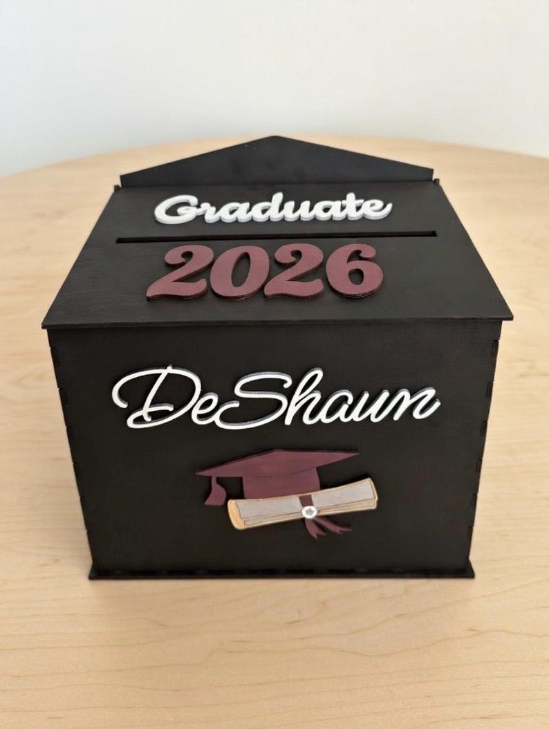 K&ouml;nnte beinhalten: Eine schwarze Holz-Abschlussbox mit einem Schlitz oben. Die Box zeigt die W&ouml;rter "Graduate 2026" und "DeShaun" in wei&szlig;er und weinroter Schrift sowie eine Illustration einer Abschlusskappe und eines Diploms.