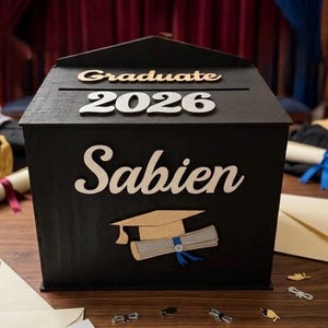 K&ouml;nnte beinhalten: Eine schwarze Holz-Abschlussbox mit den Worten "Graduate 2026" und "Sabien" in wei&szlig;er Schrift. Eine Abschlusskappe und ein Diplom sind ebenfalls abgebildet. Die Box steht auf einer Holzoberfl&auml;che mit Konfetti zum Thema Abschluss.